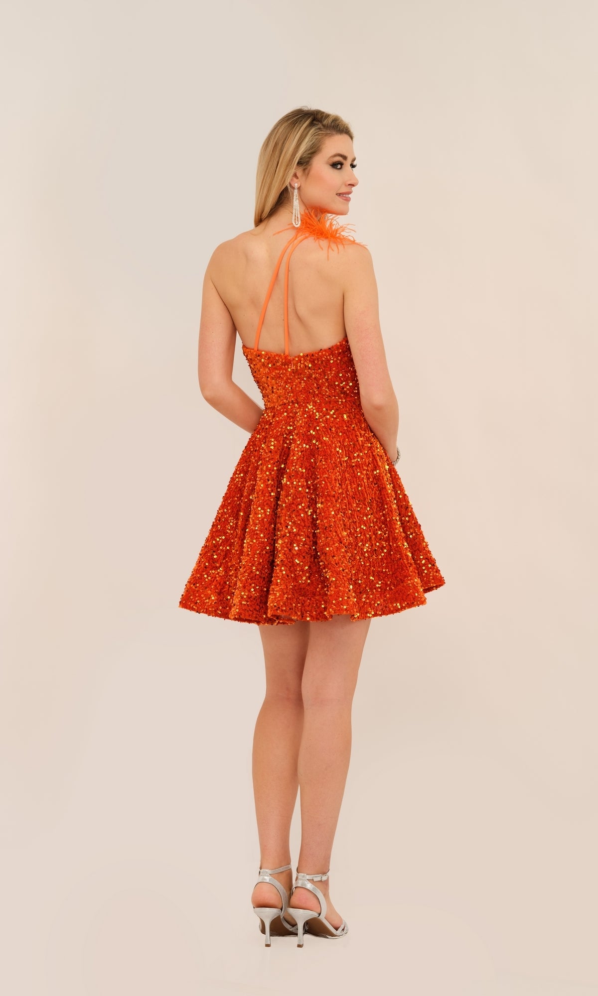 A woman in the Dave and Johnny Short Sequin Hoco Dress 11189, featuring an open back, stands in silver heels facing slightly away and looking over her shoulder, exuding playful elegance with its shimmering sequins.